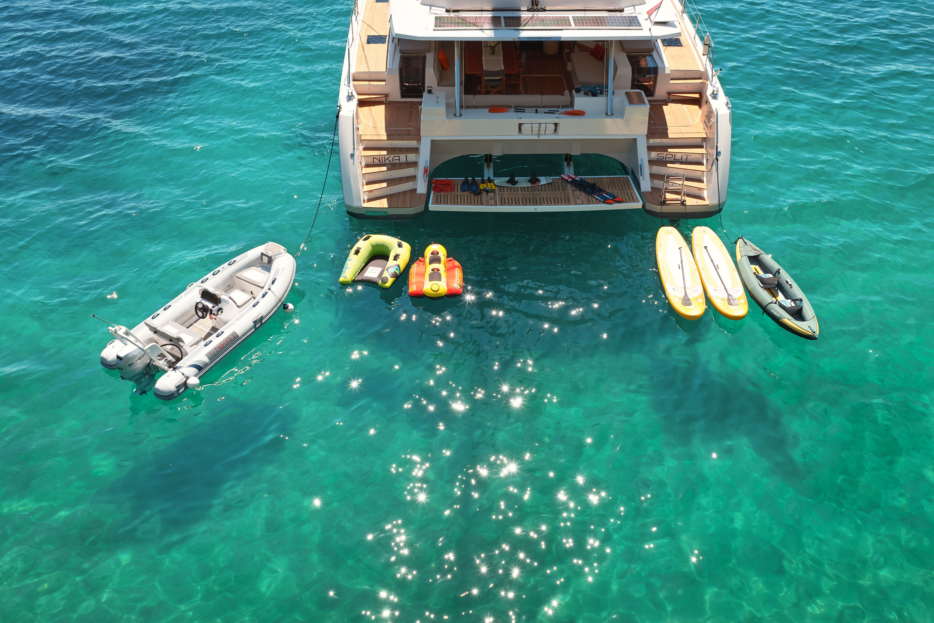 Aerial stern close, full water sports spread -- tender, banana, SUPs, kayak, clear water, NIKA stern