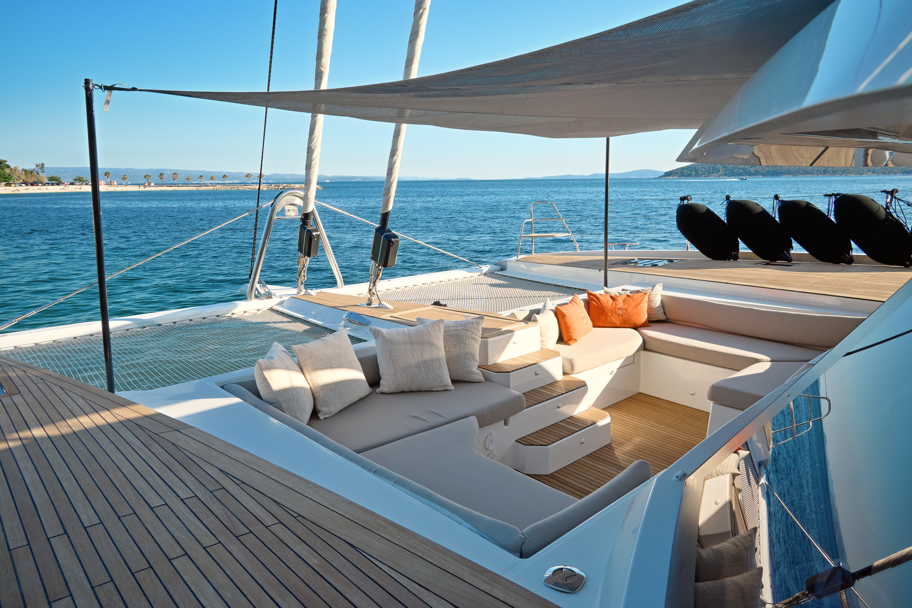 Clean foredeck lounge under sail awning, cushions arranged, open sea and island coastline beyond